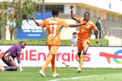 Orange-clad soccer players celebrate a goal as one raises a fist while a teammate approaches; a purple-clad goalkeeper kneels on the turf nearby.