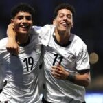 Two soccer players in white jerseys celebrate with their arms around each other on the field smiling broadly  trybalnews