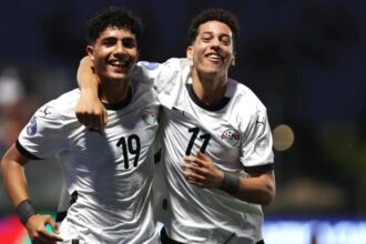 Two soccer players in white jerseys celebrate with their arms around each other on the field, smiling broadly.
