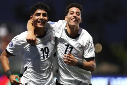 Two soccer players in white jerseys celebrate with their arms around each other on the field, smiling broadly.