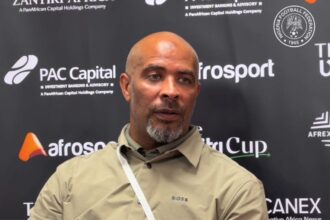 Eric Chelle in a beige shirt sits at a press backdrop, leaning backwards during a post match interview.