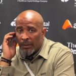 Eric Chelle in a beige shirt sits at a press backdrop touching his ear with his left hand during a post match interview  trybalnews