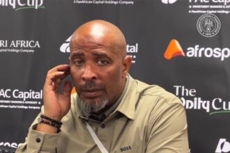 Eric Chelle in a beige shirt sits at a press backdrop, touching his ear with his left hand during a post match interview.