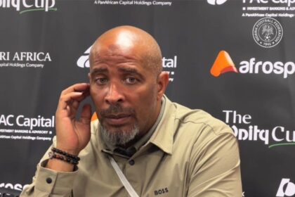Eric Chelle in a beige shirt sits at a press backdrop, touching his ear with his left hand during a post match interview.