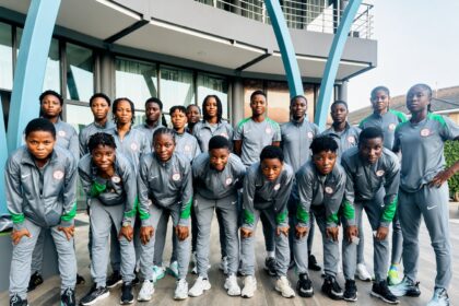 Team of young athletes in gray and green tracksuits posing for a group photo outdoors in front of a modern building with blue arches outside.