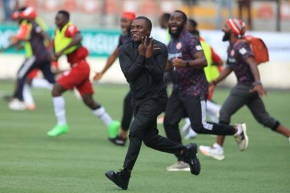 Man in black athletic wear sprints across a soccer field as teammates celebrate in the background.