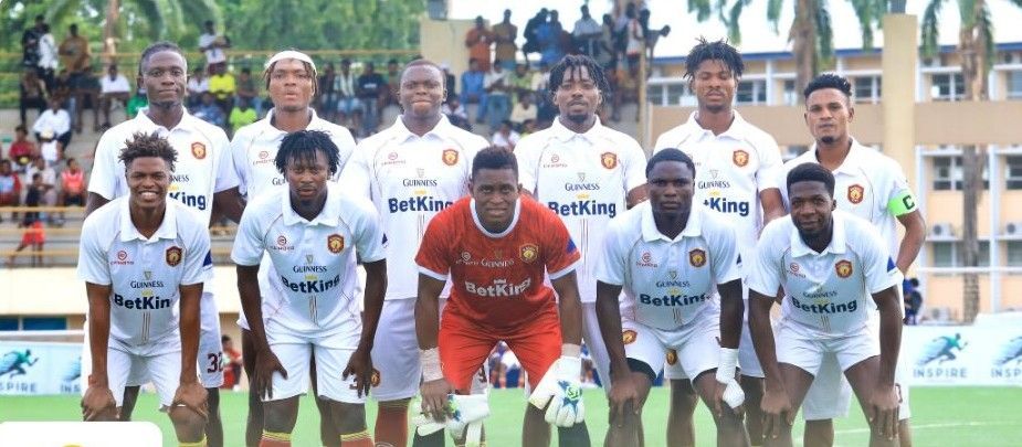 IkoroduCity Trybal News A soccer team photo with 11 players in white jerseys and a red clad goalkeeper kneeling in front posing on a field before a match trybalnews