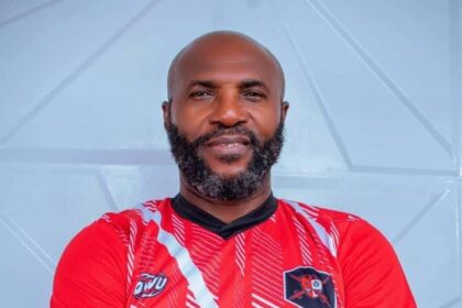 Man with a beard wearing a red sports jersey, arms crossed, standing against a light blue wall.