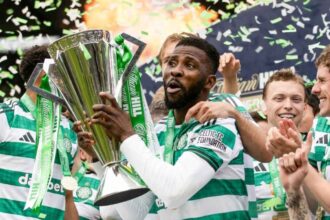 Celtic FC players celebrate victory, lifting a tall silver trophy with green ribbons and confetti flying around.