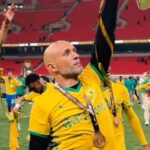 Bald man in a yellow jersey raises his arm in celebration on a football field medal hanging from his neck with teammates and red stadium seats in the background  trybalnews