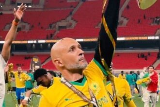 Bald man in a yellow jersey raises his arm in celebration on a football field, medal hanging from his neck, with teammates and red stadium seats in the background.