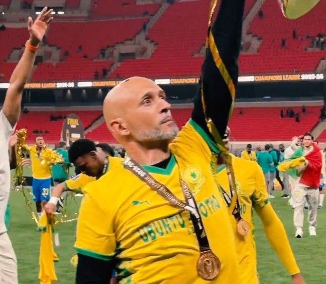 Bald man in a yellow jersey raises his arm in celebration on a football field medal hanging from his neck with teammates and red stadium seats in the background  trybalnews