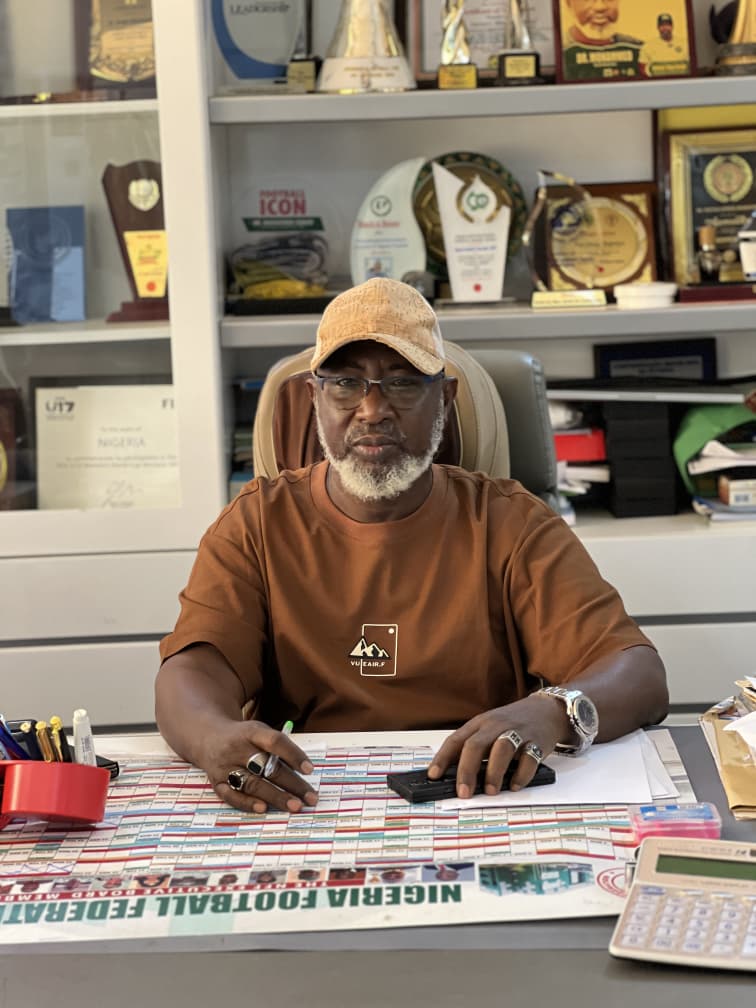 Man with a tan cap and brown shirt sits at a desk signing papers with a calculator nearby and trophies on display behind him  trybalnews