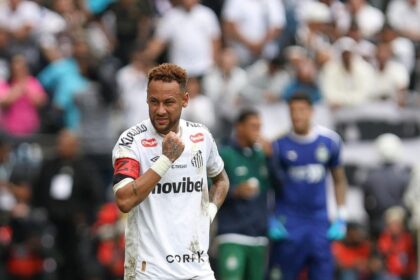 Soccer player in a white kit with a red captain's armband on his left arm, standing on a muddy field as fans cheer in the blurred stands behind him.