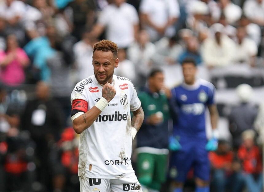 Soccer player in a white kit with a red captains armband on his left arm standing on a muddy field as fans cheer in the blurred stands behind him  trybalnews