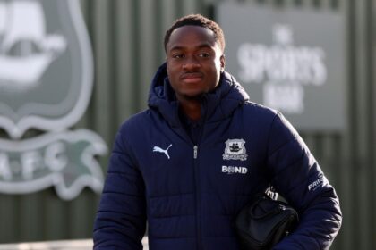 Young man in a navy puffer jacket with a crest and a Puma logo, carrying a small black bag outdoors.