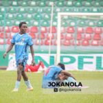Two soccer players in blue kits on a grassy field one stands while another sits on the ground near a goal stadium seating in the background  trybalnews