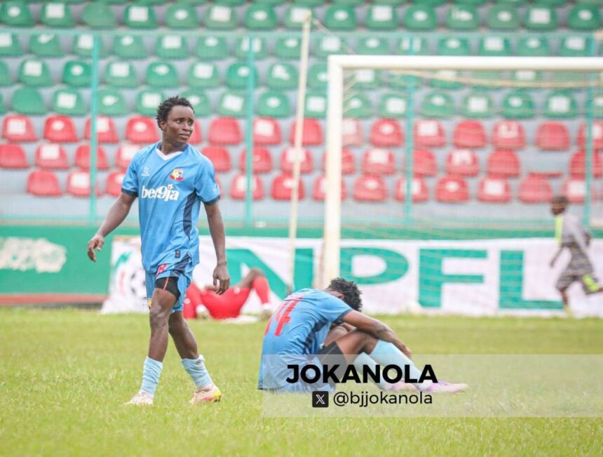 Two soccer players in blue kits on a grassy field one stands while another sits on the ground near a goal stadium seating in the background  trybalnews