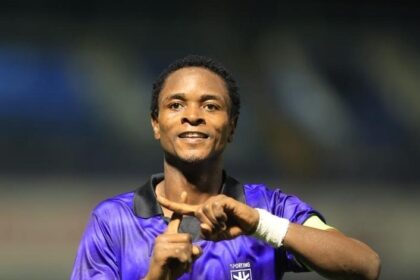 Soccer player in a purple jersey on a stadium field, smiling and making a hand gesture with both hands to celebrate the moment.