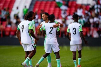 Group of soccer players in white and green uniforms celebrate on a green pitch, with numbers 9, 2, and 3 on their backs.