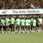 Soccer players in neon green shirts form a huddle on the training field beneath a sign reading The Artc Stadium  trybalnews