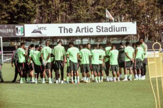 Soccer players in neon green shirts form a huddle on the training field beneath a sign reading The Artc Stadium.