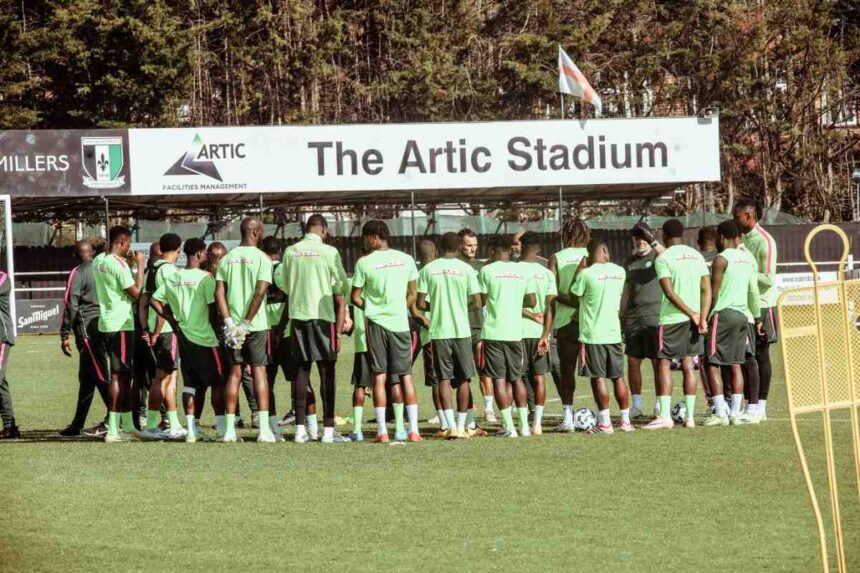 Soccer players in neon green shirts form a huddle on the training field beneath a sign reading The Artc Stadium  trybalnews