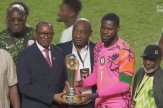 Group of men on a soccer field presenting a trophy to a player in a bright pink jersey/kit.