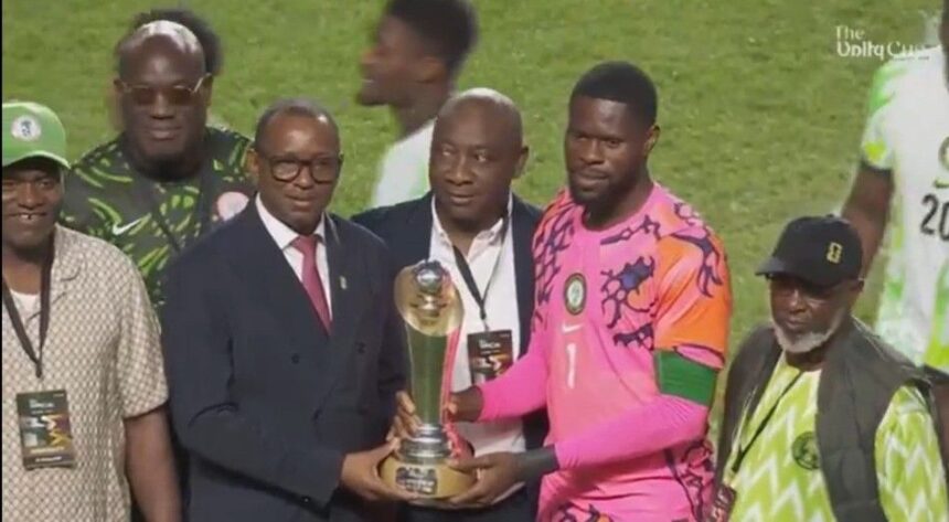 Group of men on a soccer field presenting a trophy to a player in a bright pink jerseykit  trybalnews