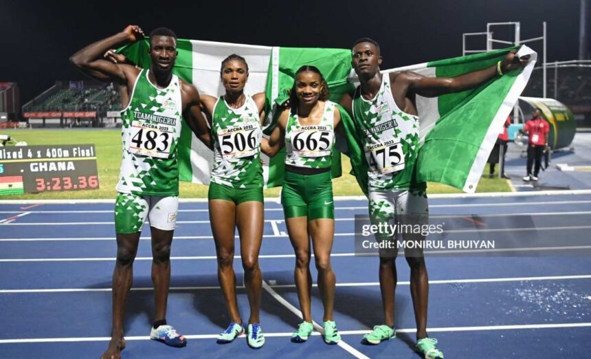 Four Nigerian athletes on a track celebrate a win holding a green and white flag behind them wearing Team Nigeria uniforms with bib numbers 483 506 665 and 475  trybalnews