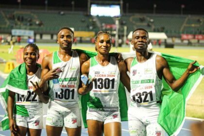 Four Nigerian track athletes pose together after a race, holding a green-and-white Nigerian flag on a stadium track.
