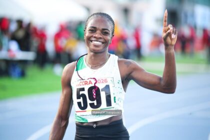 Smiling female long-distance runner on a track, wearing bib 591 with Accra 2026, raising one finger in victory.
