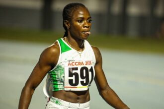 Female sprinter in a white and green athletic outfit with bib 'ACCRA 2026' and number 591, walking on a track after a race.