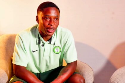 Young man in a mint green polo with a Nigeria Football Federation crest sitting in a beige armchair in a softly lit room.