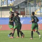 Soccer players in green uniforms celebrate on a grass field embracing each other after a goal as stadium seats loom in the background  trybalnews