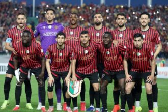 Soccer team in red-and-black striped jerseys posing for a group photo on the field; goalkeeper in purple stands behind them.