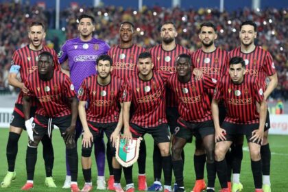 Soccer team in red-and-black striped jerseys posing for a group photo on the field; goalkeeper in purple stands behind them.