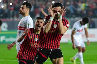 Two soccer players in red-and-black striped jerseys celebrate on the field, one making a playful 'glasses' gesture with his hands around his eyes.