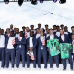 Group of men in navy suits posing on a stage outdoors some wear red and white scarves two players hold green jerseys numbered 24 and 25  trybalnews