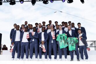 Group of men in navy suits posing on a stage outdoors; some wear red-and-white scarves, two players hold green jerseys numbered 24 and 25.
