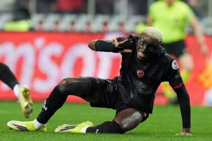 Soccer player in black kit sliding on grass, reaching with one hand during a match, smiling.