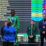 Winners podium ceremony a central athlete in green with a medal stands on the first place platform flanked by others in blue and dark outfits as a large scoreboard scrolling names and years looms behind them  trybalnews