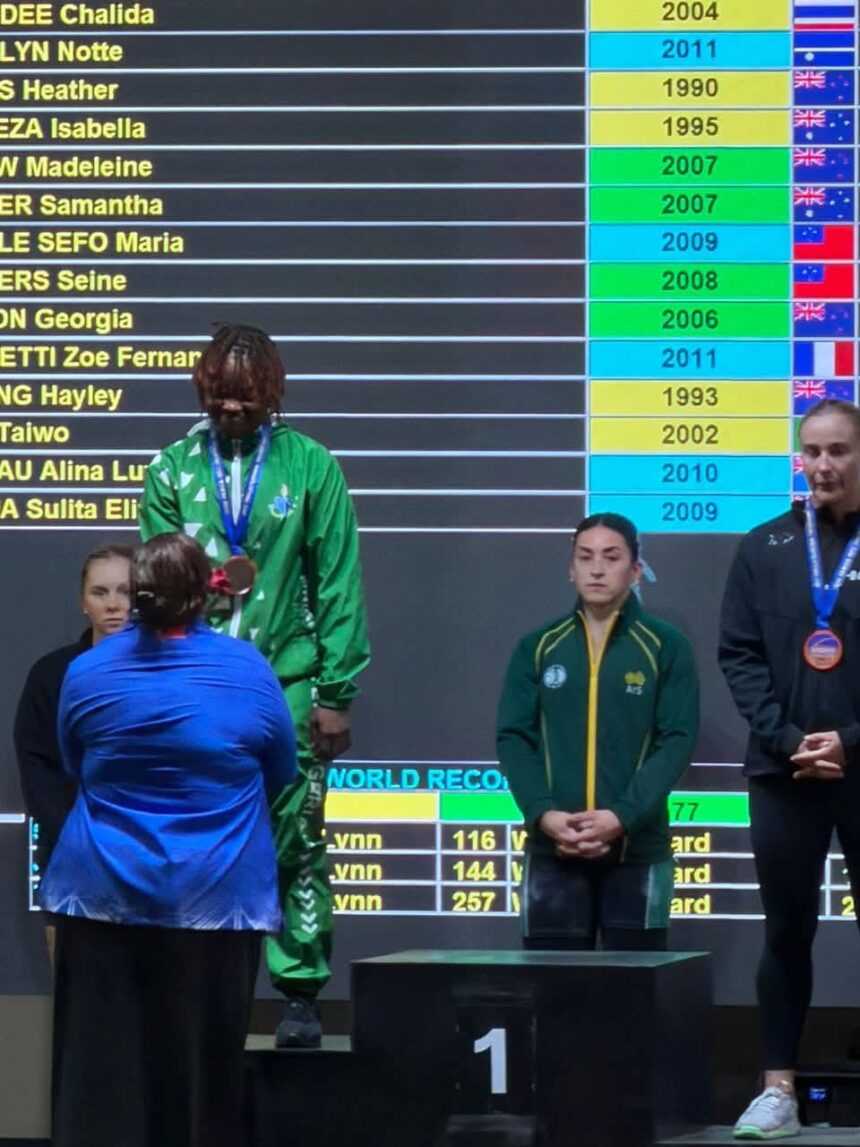 Winners podium ceremony a central athlete in green with a medal stands on the first place platform flanked by others in blue and dark outfits as a large scoreboard scrolling names and years looms behind them  trybalnews