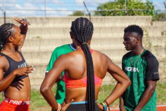 Athletes on a track field chat; central figure is a woman with braided hair seen from behind.