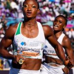 Two female sprinters race toward the finish in a crowded stadium foreground runner in a white and green kit holds a baton as she moves forward  trybalnews