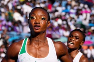 Two female sprinters race toward the finish in a crowded stadium; foreground runner in a white-and-green kit holds a baton as she moves forward.