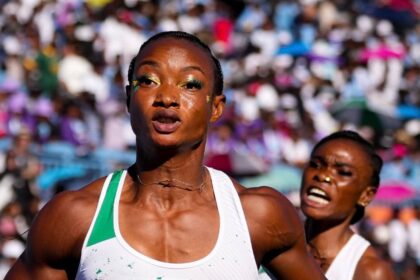 Two female sprinters race toward the finish in a crowded stadium; foreground runner in a white-and-green kit holds a baton as she moves forward.
