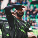 Soccer player in a black and green striped jersey shields his eyes with his hand while looking upward stadium crowd in the background  trybalnews