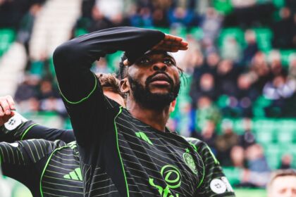 Soccer player in a black and green striped jersey shields his eyes with his hand while looking upward, stadium crowd in the background.