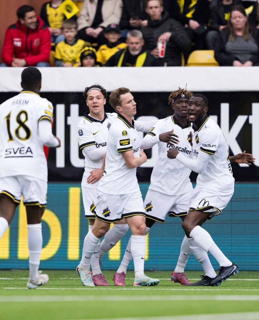 WhatsApp Image 20260503 at 180434 Trybal News Soccer players in white and yellow kits celebrate a goal on the field teammates embracing near the sideline while fans watch from the stands trybalnews